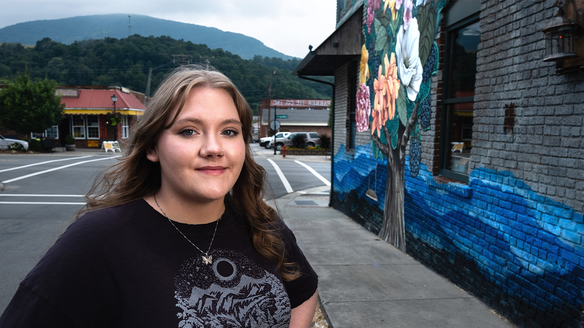 A woman posing for a portrait on a street in a mountainous town with a painted building mural to her right.