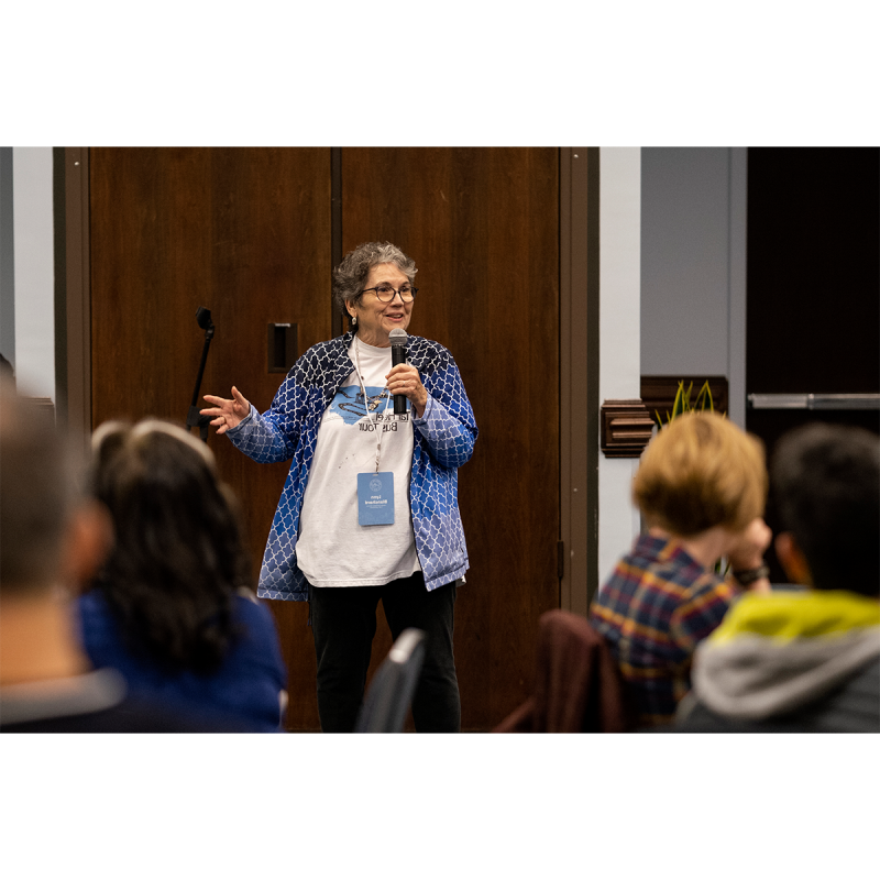 一个女人, named 林恩·布兰查德, holding a microphone and speaking to a group of bus tour attendees in an indoor meeting room.