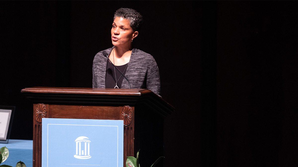 A woman, Michelle Alexander, speaking from a podium on a stage at a lecture.