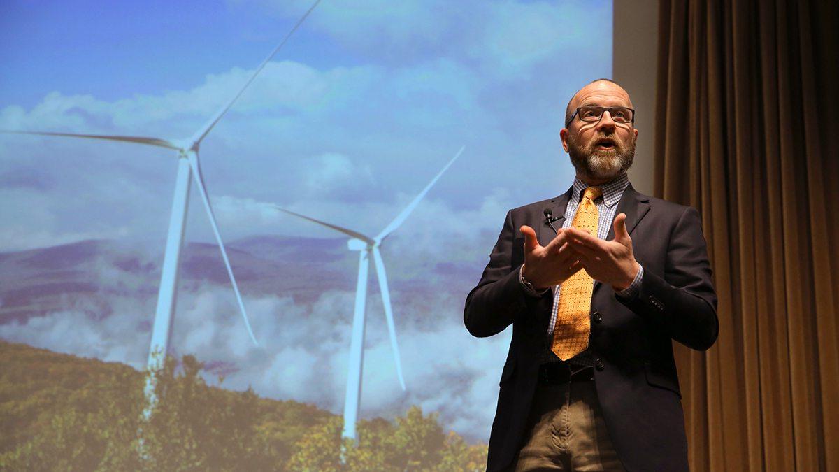 Man wearing microphone and speaking in front of powerpoint image with windmills.