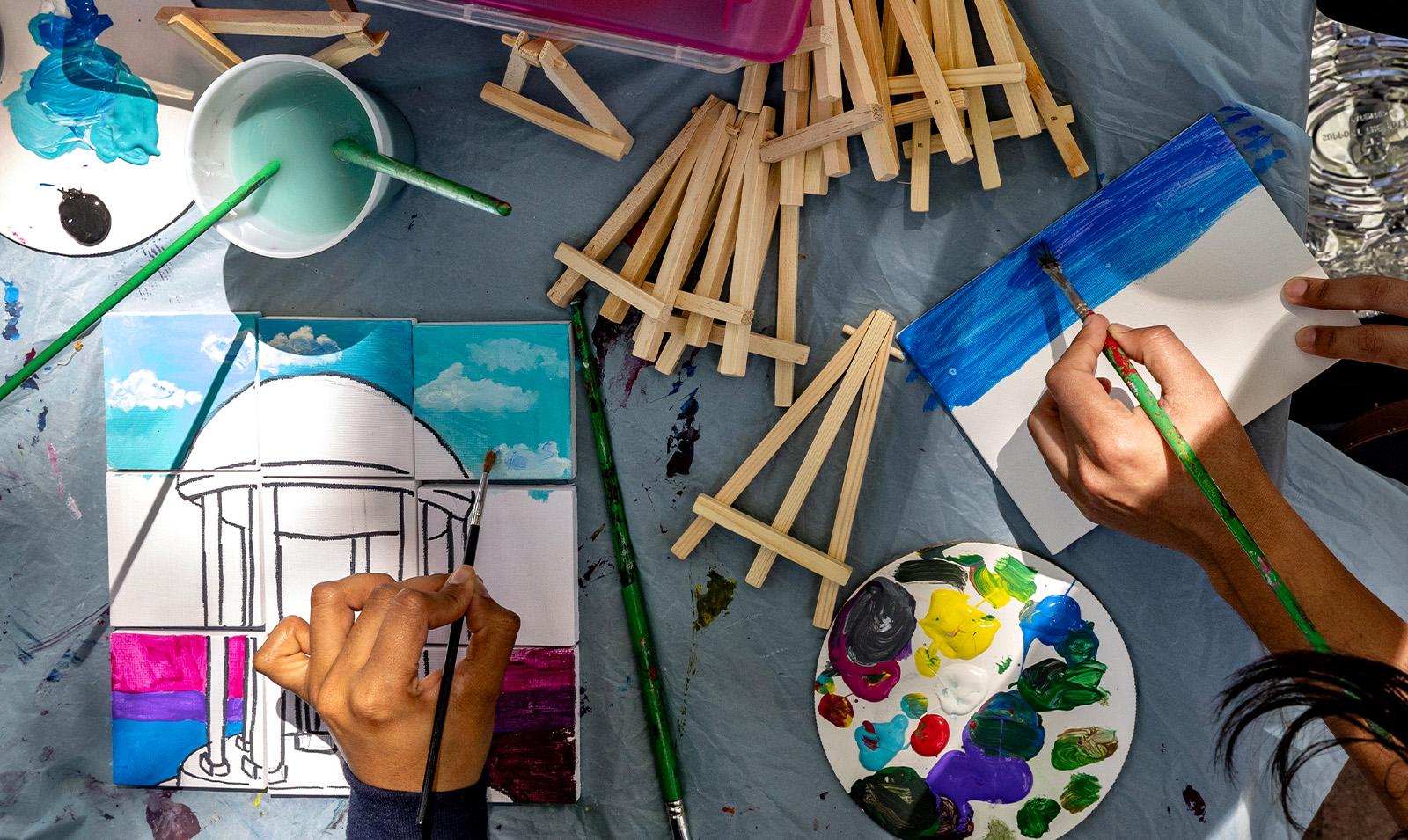 Overhead image with two people's hands seen finishing paintings, one of which is of the Old Well.